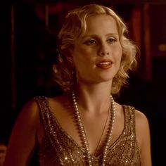 a blonde woman in a gold dress with pearls on her neck and chest, looking at the camera