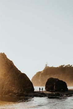 two people standing on the beach next to large rocks