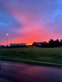 the sun is setting over a grassy field with buildings in the distance and cars driving down the road