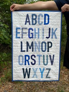 a person holding up a quilted alphabet sign in front of some trees and bushes