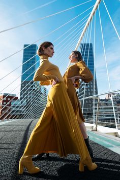 two women in yellow dresses are standing on a bridge and looking at each other with city buildings in the background