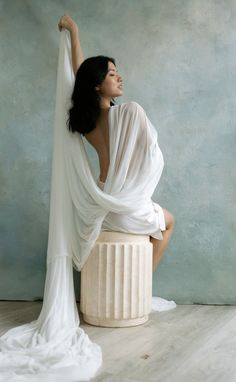 a woman sitting on top of a white pillar in front of a blue wall wearing a white dress