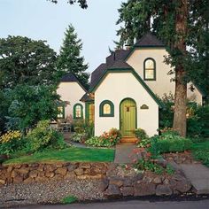 a white house with green trim surrounded by trees and flowers in the front yard area