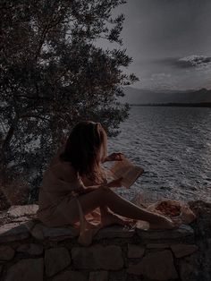 a woman sitting on the edge of a cliff reading a book by the water's edge