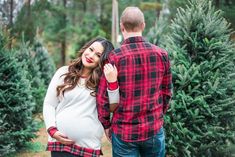 a pregnant woman standing next to a man in front of christmas tree trees with her hand on her belly