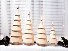 three wooden christmas trees on a table with pine cones in the foreground and white curtains behind them