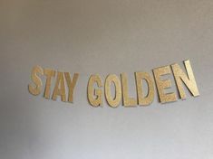 the word stay golden is cut out from gold foil on a white wall and hung by a string