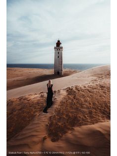 a woman standing in the sand next to a light house on top of a hill