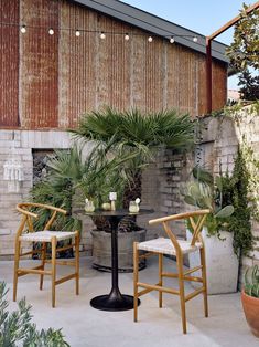 two chairs and a table in front of a brick wall with potted plants on it