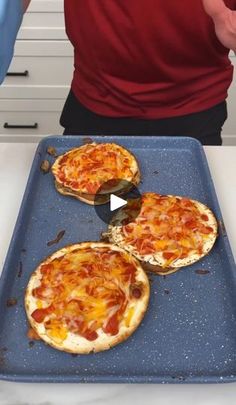 three pizzas sitting on top of a blue pan next to a person in a red shirt