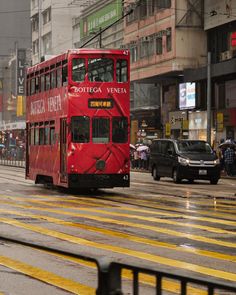 a red double decker bus driving down a street next to tall buildings and people walking on the sidewalk