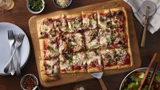 a pizza sitting on top of a wooden cutting board next to bowls of salad and utensils