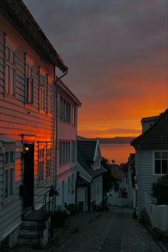 the sun is setting over some houses and buildings on the side of the road with water in the distance