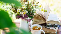 an open book and glasses on a table next to a cup of tea, with flowers in the background