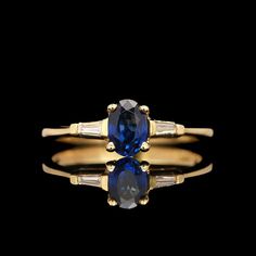 a gold ring with an oval blue sapphire and baguettes on the sides, set against a black background