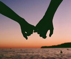 two people holding hands while standing in front of the ocean at sunset with boats in the background