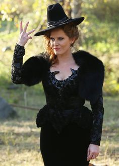 a woman in a black dress and hat with her hand up to the side while standing outside