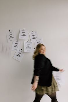 a woman in black jacket and skirt standing next to wall with white papers on it