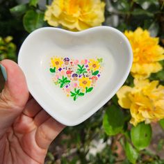 a hand holding a white heart shaped dish with flowers on it in front of yellow roses