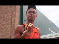 a man holding a hot dog in front of a brick building and wearing an orange tank top