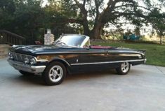 an old black convertible car parked in front of a driveway with trees and bushes behind it