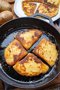 four pieces of food cooking in a skillet on a wooden table next to potatoes