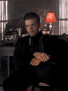 a man in a black suit sitting on a chair next to a table and lamp