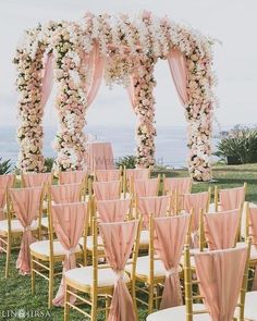 an outdoor ceremony setup with pink and white flowers on the arch, gold chairs and draping