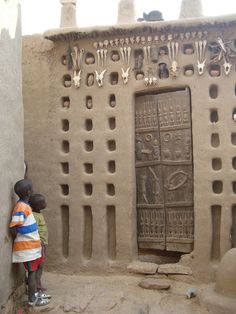 two children are standing in front of a mud house with carvings on the walls and doors