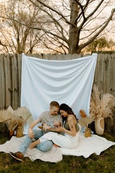 a man and woman sitting on top of a blanket in the grass next to a fence