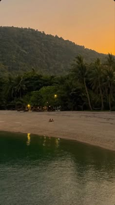 people are sitting on the beach at sunset with mountains in the background and trees lining the shore