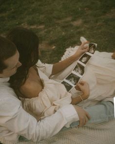 a man and woman are sitting on the grass together, holding an old photo album