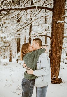 a man and woman are kissing in the snow near some tall trees with their arms around each other