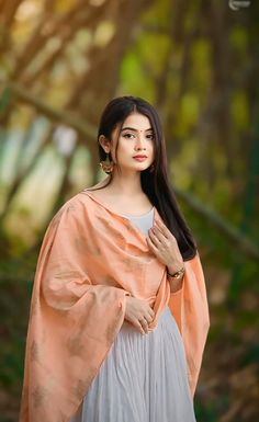 a woman with long black hair wearing an orange shawl and white dress standing in front of trees