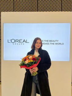 a woman standing in front of a large screen holding a bouquet of flowers and smiling at the camera