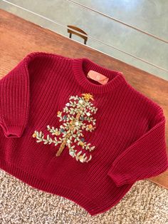 a red sweater with an embroidered christmas tree on the front and bottom, sitting on carpet