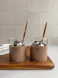 two glasses filled with chocolate pudding and whipped cream on a wooden tray next to a white tile wall