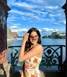 a woman in a floral dress is posing for a photo by the water with her sunglasses on