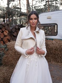 a woman in a white dress and jacket standing next to a pile of hay with a camper behind her