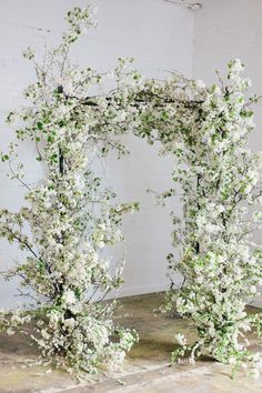 an arch made out of branches with white flowers and greenery on the sides, in front of a brick wall