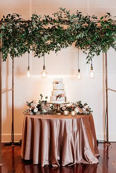 a wedding cake on top of a table with greenery and lights hanging from the ceiling