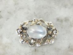 "This is an interesting and unique brooch from the 1930's depicting a classic motif of rosettes and ivy leaves. The leaves ornately fill this piece making a classic image for this period. We've set this vintage brooch with an oval moonstone gem, one with excellent clarity and plenty of light! Love this piece but want to make it more versatile, we here at Market Square Jewelers along with our master metalsmiths can convert this fantastic brooch into a necklace or pendant for an additional fee, pl Vintage Silver Brooch With Gemstone, Vintage Silver Gemstone Brooch, Art Nouveau Cabochon Brooches For Weddings, Art Nouveau Cabochon Wedding Brooches, Heirloom Silver Brooch With Rose Cut Diamonds, Heirloom Silver Brooches With Rose Cut Diamonds, Vintage Silver Brooch With Rose Cut Diamonds, Vintage Silver Brooches With Rose Cut Diamonds, White Vintage Cabochon Brooches