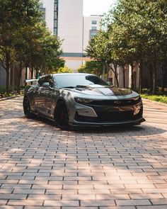 a gray sports car parked in front of trees on a brick road with buildings in the background