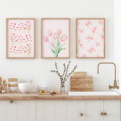 three framed pictures on the wall above a kitchen counter with flowers and other items in vases