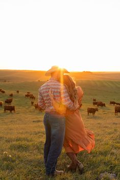 a man and woman standing in front of cows on a grassy field at sundown