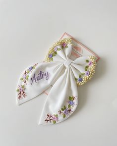 an embroidered white bow with flowers and pearls on the side that says,'autry '