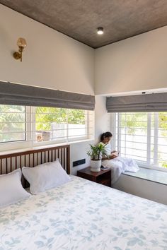 a person sitting on a bed in a room with two windows and a plant next to the bed