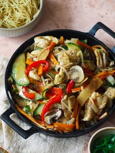 a skillet filled with noodles and vegetables on top of a table next to a bowl