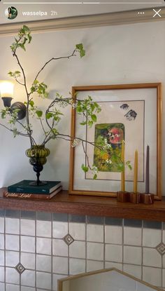 a vase filled with green plants sitting on top of a mantle next to a framed painting