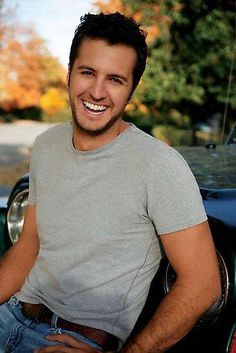 a smiling man standing next to a parked car in front of a tree filled street